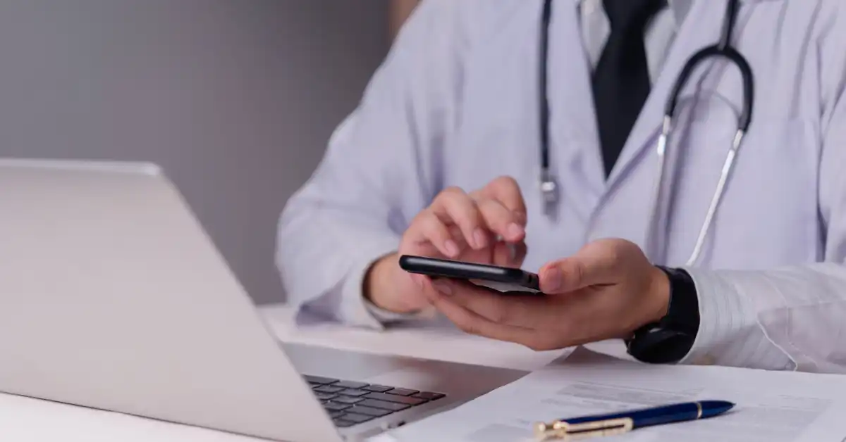 Doctor using a smartphone beside a laptop while managing telehealth consultation tasks.