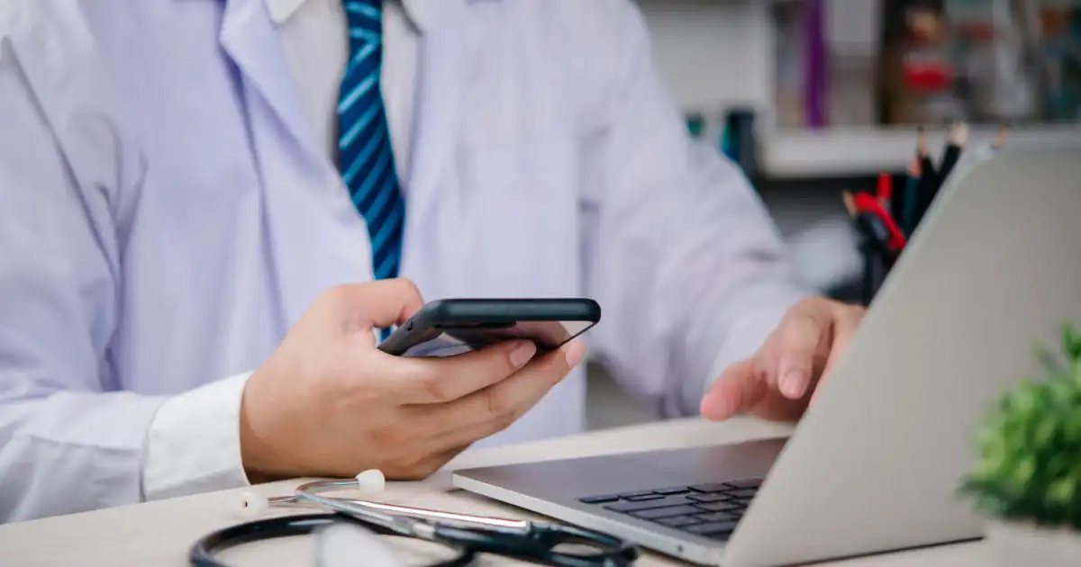 Doctor using smartphone while working on laptop during telehealth consultation.